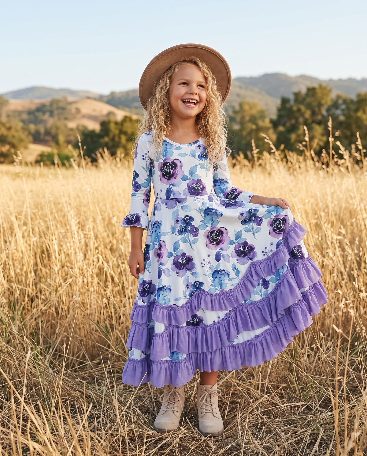A young girl smiling in a golden field, wearing a white tiered maxi dress with a purple and blue floral print, a brown wide-brim hat, and beige boots.
